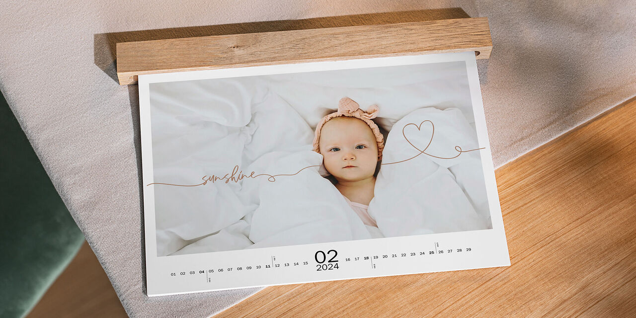 Auf einem Tisch aus Holz liegt ein Wandkalender mit Babymotiven im Querformat. Zu sehen ist die Seite für den Februar, eine Holzleiste verdeckt die Ringbindung.