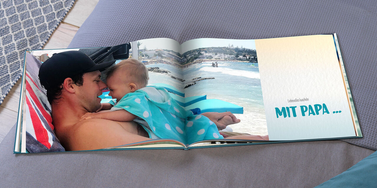 Ein aufgeschlagenes CEWE FOTOBUCH zeigt Evan am Strand, seine Tochter Hannah liegt auf ihm. Rechts im Buch steht "Liebevolles kuscheln MIT PAPA...".