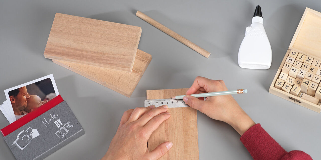 Eine Person bastelt mit Holz und Material auf einem Tisch. Es liegen zwei Holzplatten, ein Bleistift, ein Zirkel und eine Flasche Kleber auf dem Tisch. Neben der Person steht ein CEWE Fotobuch.