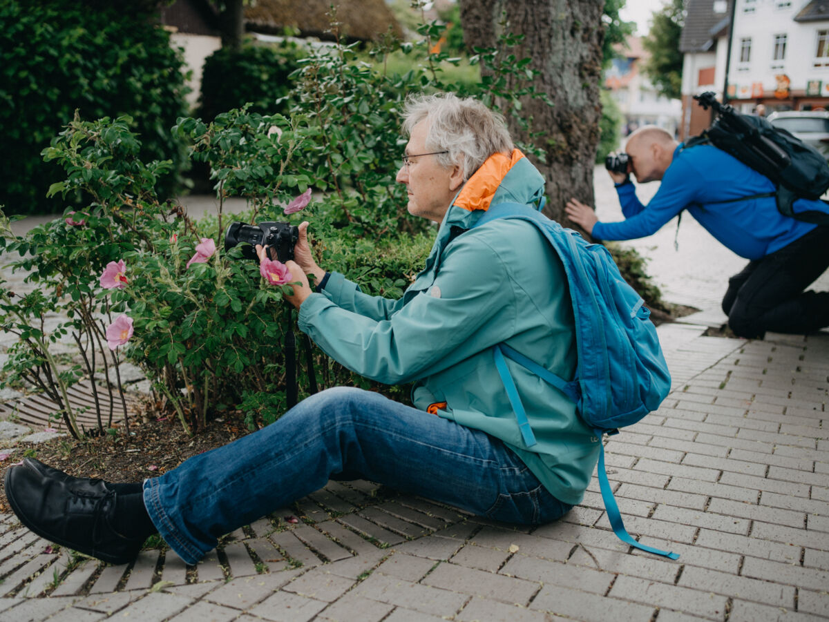 Zwei Personen fotografieren Blumen am Straßenrand, eine sitzt auf dem Gehweg mit Rucksack, die andere kniet im Hintergrund.