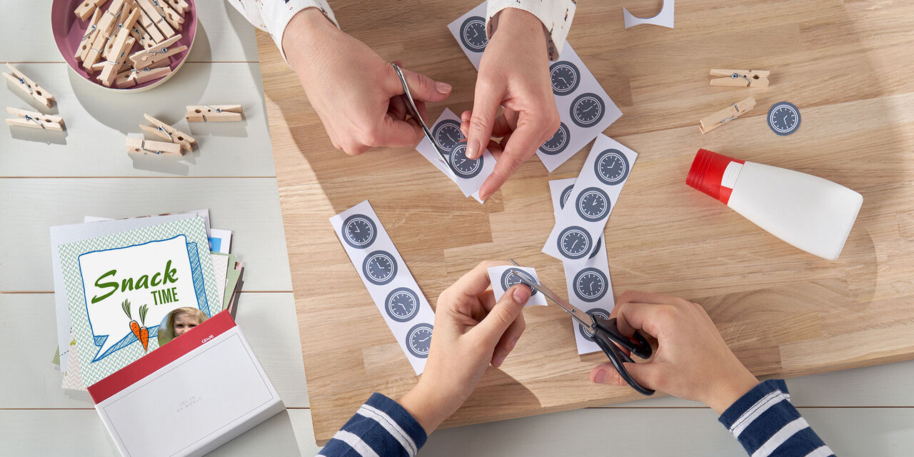 Zwei Personen schneiden bunte Papierformen aus, die eine kreative Bastelzeit zeigen. Auf dem Tisch befinden sich eine Schere und Kleber.