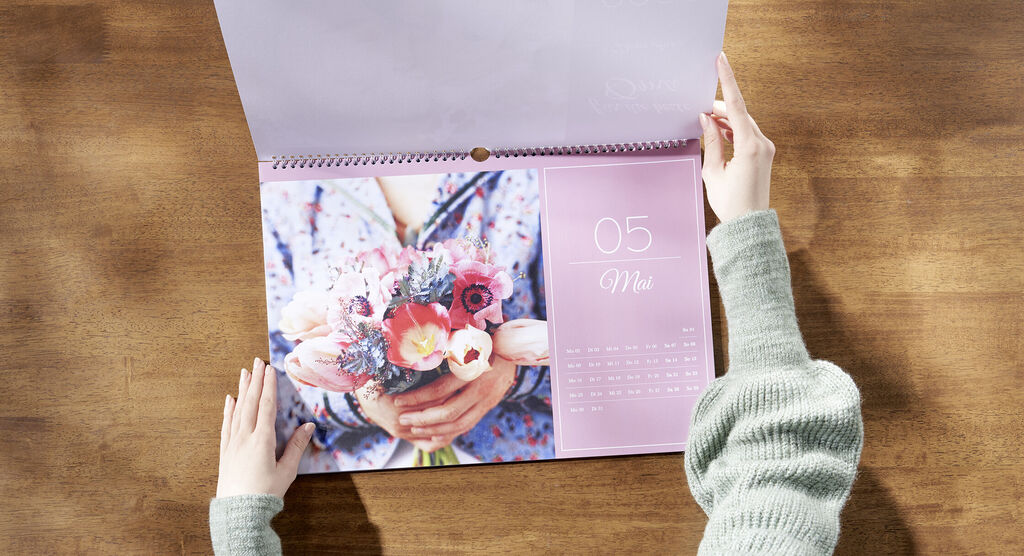 Hände halten einen Kalender auf einem Holztisch, der einen Monat mit blühenden Blumen zeigt