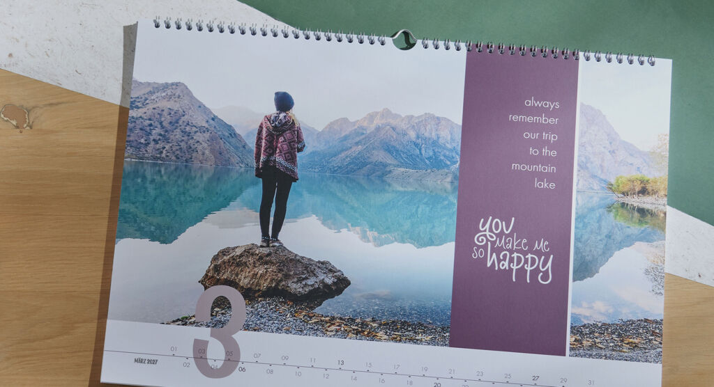 Eine Person steht auf einem Stein am Ufer eines klaren Bergsees. Im Hintergrund sind Berge, die den Himmel spiegeln. Der Kalender zeigt den Monat März an und hat einen positiven Spruch.